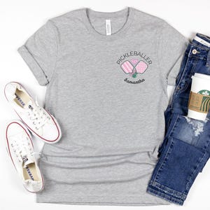 Pu&ograve; includere: Una maglietta grigio melange con un piccolo disegno ricamato con la scritta "Pickleballer" con due racchette da pickleball e un cuore. Il nome "Samantha" &egrave; sotto il disegno. Scarpe da ginnastica bianche e jeans blu sono anche nell'immagine.
