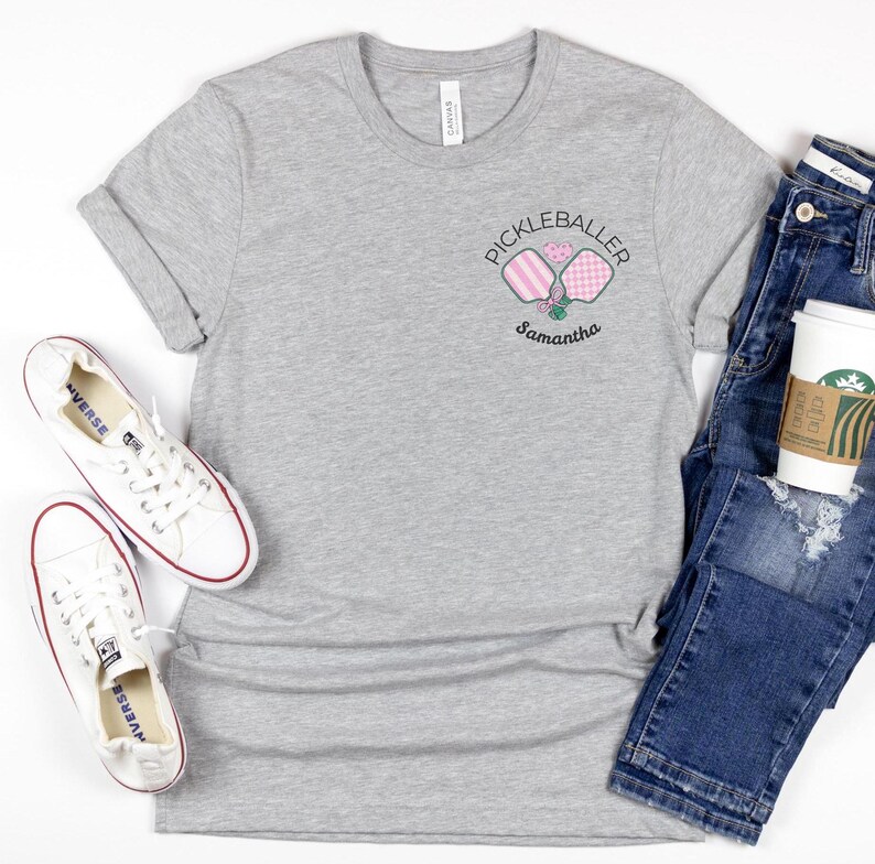 Pu&ograve; includere: Una maglietta grigio melange con un piccolo disegno ricamato con la scritta "Pickleballer" con due racchette da pickleball e un cuore. Il nome "Samantha" &egrave; sotto il disegno. Scarpe da ginnastica bianche e jeans blu sono anche nell'immagine.