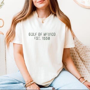 May include: A cream-colored short-sleeve t-shirt featuring the dark gray distressed text GULF OF MEXICO EST. 1550 printed across the chest. The garment has a crew neckline and a relaxed fit, paired with light blue denim jeans.
