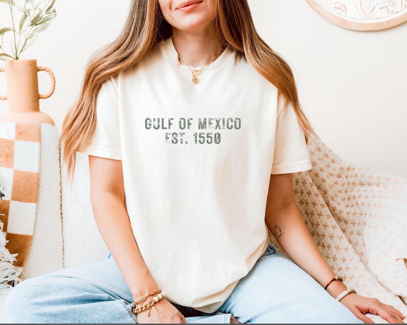 May include: A cream-colored short-sleeve t-shirt featuring the dark gray distressed text GULF OF MEXICO EST. 1550 printed across the chest. The garment has a crew neckline and a relaxed fit, paired with light blue denim jeans.
