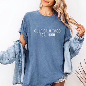 May include: A blue short-sleeved t-shirt with a crew neckline featuring the white distressed text GULF OF MEXICO EST. 1550 printed on the chest. The shirt has a relaxed fit and is worn with a light blue denim jacket draped over the shoulders.
