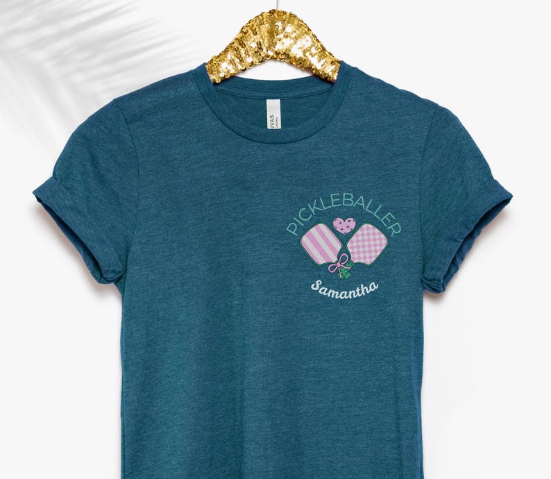 May include: Teal blue t-shirt with a white graphic of two pink and white pickleball paddles with the text "Pickleballer Samantha" in white.