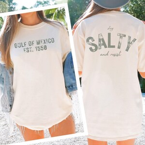 May include: An off-white short-sleeve t-shirt featuring the text GULF OF MEXICO EST. 1550 on the front and the text stay SALTY and resist on the back in a distressed green font. The shirt has a crew neckline and a relaxed, loose-fitting silhouette.