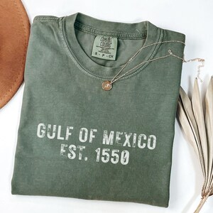 May include: An olive green short-sleeve t-shirt with a crew neckline. The shirt features the white text GULF OF MEXICO EST. 1550 printed on the front. A Comfort Colors brand label is visible at the inner neck, indicating size S, P, CH.