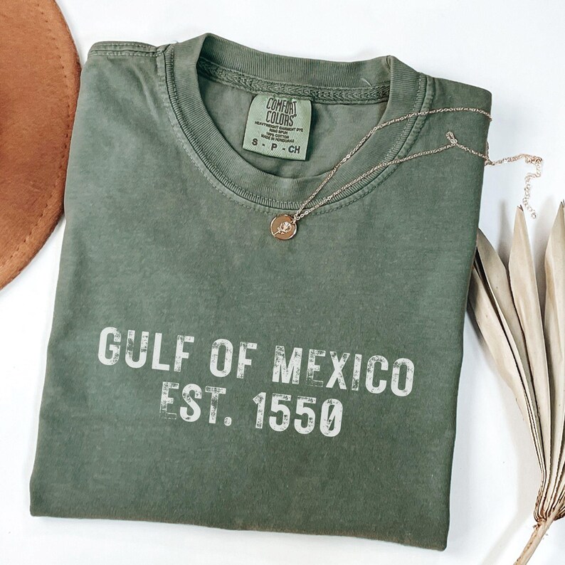 May include: An olive green short-sleeve t-shirt with a crew neckline. The shirt features the white text GULF OF MEXICO EST. 1550 printed on the front. A Comfort Colors brand label is visible at the inner neck, indicating size S, P, CH.