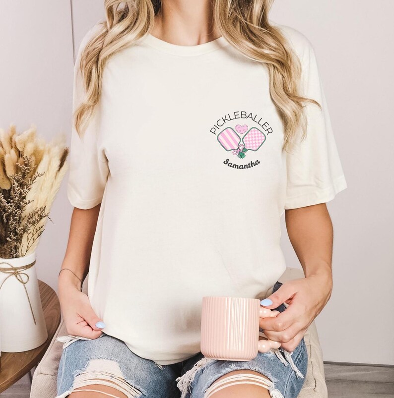 Pu&ograve; includere: T-shirt color crema con una piccola grafica di due racchette da pickleball e la scritta "Pickleballer" sopra il nome "Samantha". Le racchette sono rosa e a scacchi. La maglietta &egrave; in stile casual.