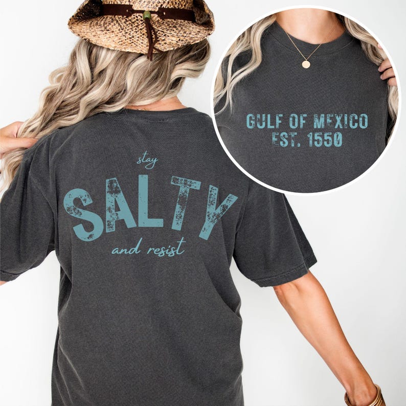 May include: A charcoal gray t-shirt featuring light blue text. The back displays the words stay SALTY and resist. The front circular inset shows the text GULF OF MEXICO EST. 1550 in a matching light blue color with a distressed, weathered texture.