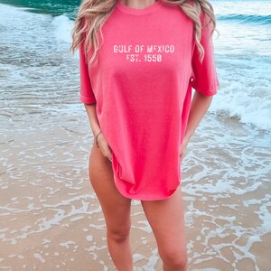 May include: A coral-colored short-sleeve t-shirt featuring the white distressed text GULF OF MEXICO EST. 1550 printed on the chest. The garment has a crew neckline and a relaxed fit, constructed from a solid-colored fabric with a soft, worn-in appearance.