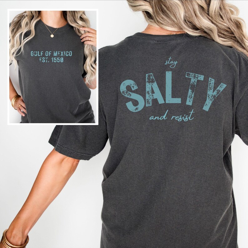 May include: A charcoal gray short-sleeve t-shirt featuring the text "GULF OF MEXICO EST. 1550" on the front chest and the large light blue distressed text "stay SALTY and resist" printed on the back of the garment.