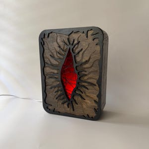 Puede incluir: Lámpara rectangular de madera con marco teñido oscuro y centro de veta de madera más clara. El centro presenta una forma orgánica roja y brillante. El diseño incluye intrincados patrones cortados con láser que se asemejan a ramas o venas.