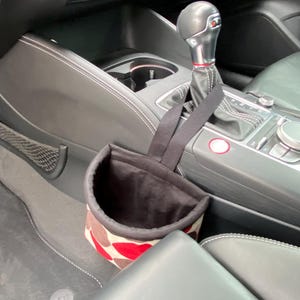 May include: A black and white patterned fabric trash can with a black strap hanging from the gear shift of a car. The trash can is hanging from the gear shift of a car.