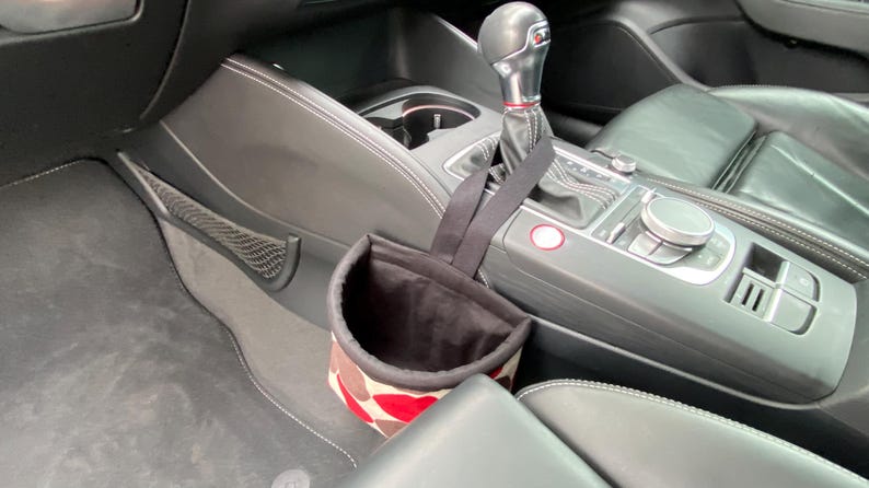 May include: A black and white patterned fabric trash can with a black strap hanging from the gear shift of a car. The trash can is hanging from the gear shift of a car.