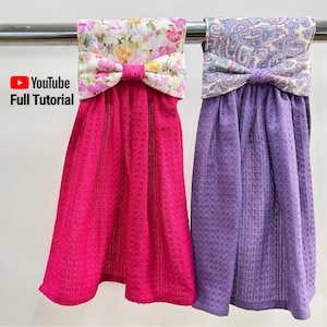 May include: Two decorative kitchen towels hanging on a metal bar. One is bright pink with a floral bow, the other is purple with a paisley bow. The towels have a textured waffle weave. The text "YouTube Full Tutorial" is visible.