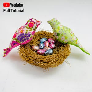 以下が含まれることがあります： 花柄の布製の鳥が2羽、チョコレートのイースターエッグが入った編まれた巣の中にいます。鳥はピンクとグリーンで白い花が咲いています。巣は茶色で自然繊維で作られています。