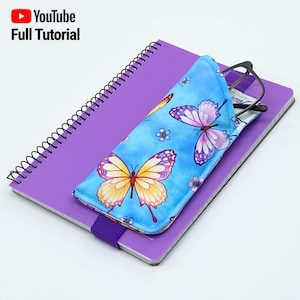 May include: A blue and yellow butterfly patterned glasses case with a purple elastic band. The case is sitting on top of a purple notebook with a spiral binding.