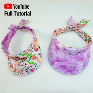 May include: Two fabric visors. One visor has a white background with a floral pattern of pink, red, and green flowers. The other visor has a purple marbled pattern. Both visors have a matching tie.
