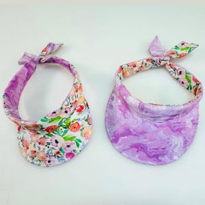 May include: Two sun visors, one with a floral print and the other with a purple marbled print. Both visors have a tie at the back.