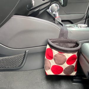 May include: A red and brown polka dot trash can with a black liner hanging from a hook in a car.