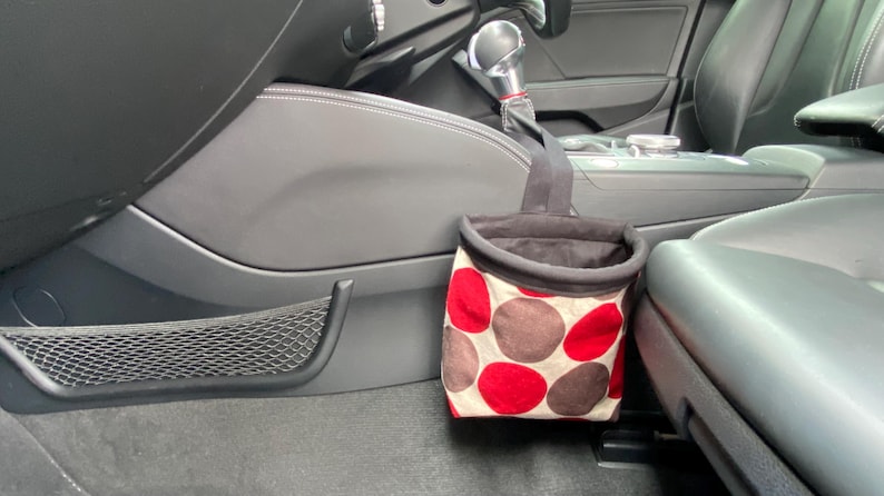 May include: A red and brown polka dot trash can with a black liner hanging from a hook in a car.