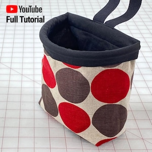 以下が含まれることがあります： 黒の布地裏地と赤と茶色の水玉模様が付いた、ベージュの小さな布地バッグ。バッグには黒い布地のハンドルが付いています。