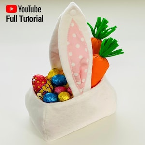 May include: White bunny-shaped Easter basket with pink polka dot ears, filled with chocolate eggs and carrot-shaped decorations.  The basket has a button closure on the top.