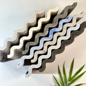 May include: A selection of wavy, textured decorative tiles in shades of grey, white, and blue. The tiles are arranged diagonally, highlighting their unique, undulating design. Some tiles have flecks of colour. The tiles are likely made of concrete.