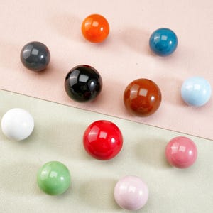 Könnte beinhalten: Eine Sammlung von elf glänzenden, kugelförmigen Knöpfen in verschiedenen Farben, darunter Orange, Türkis, Schwarz, Braun, Rot, Rosa, Grün, Weiß und Hellblau. Die Knöpfe sind auf einer zweifarbigen rosa und grünen Oberfläche angeordnet.