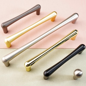 Könnte beinhalten: Eine Auswahl an modernen Schrankgriffen in verschiedenen metallischen Ausführungen. Die Auswahl umfasst mehrere Griffstangen in Bronze, Gold, Silber und Schwarz sowie einen passenden Knopf. Präsentiert auf einem zweifarbigen rosa-grünen Hintergrund.