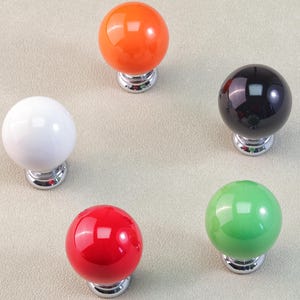 Peut inclure: Cinq boutons de meuble color&eacute;s dispos&eacute;s en cercle. Les boutons sont brillants et sph&eacute;riques, avec des couleurs comprenant le blanc, l'orange, le noir, le rouge et le vert. Chaque bouton a une base argent&eacute;e.