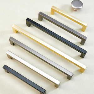 May include: A collection of modern cabinet hardware, including several rectangular bar pulls in gold, black, silver, and gray finishes. A matching round knob is also included, all on a light background.