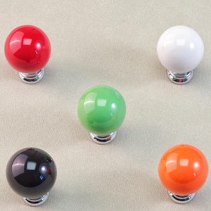 May include: Five colorful cabinet knobs in red, white, green, black, and orange. Each knob is a glossy sphere mounted on a silver-toned base. The knobs are arranged on a neutral, light-colored surface.