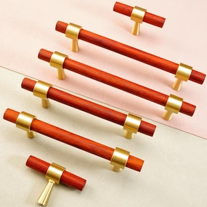May include: A collection of wooden cabinet pulls with a reddish-brown finish and gold-toned metal accents. The pulls are arranged on a two-tone pink and cream background, showcasing their design and hardware details.