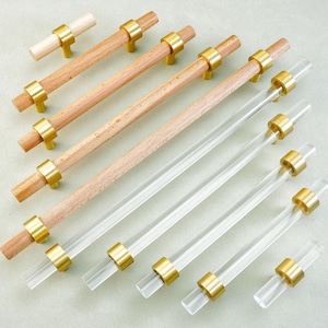 May include: A collection of cabinet hardware, including wooden and clear acrylic pulls with brass-colored accents. The wooden pulls are light brown, while the acrylic pulls are transparent. The hardware is arranged on a light green surface.