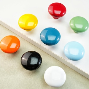 May include: A collection of eight colorful cabinet knobs in various glossy finishes. The knobs are arranged on a white surface and include shades of red, yellow, orange, green, blue, black, and white. These decorative knobs are ideal for adding a pop of color.