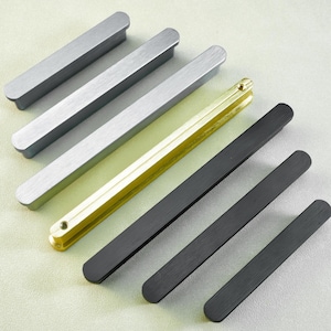 May include: An assortment of cabinet pulls in various metallic finishes. The collection includes silver, gold, and black rectangular bar-shaped handles. The pulls are arranged diagonally on a light gray surface, showcasing their different colors and designs.