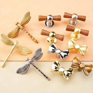 Puede incluir: Una colección de tiradores y pomos decorativos para armarios en varios diseños. La selección incluye estilos de libélula, lazo y barra de madera. Los acabados incluyen tonos dorados, plateados y bronce. Los pomos están sobre un fondo neutro.