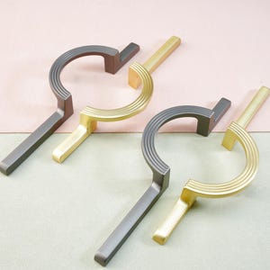 Pode incluir: Quatro puxadores de armário com design moderno. Dois são cinza escuro e dois dourados. Cada puxador tem uma alça curva com textura canelada e uma base reta e retangular. Os puxadores são dispostos em um fundo bicolor rosa e verde.