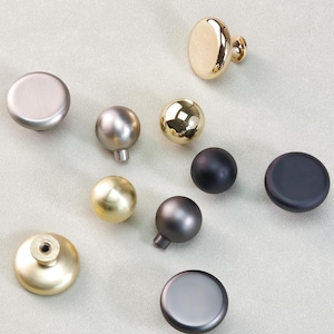 May include: An assortment of cabinet knobs in various metallic finishes, including gold, silver, and black. The knobs are round and oval, with a mix of matte and shiny surfaces. The hardware is displayed on a light gray background.