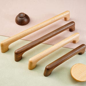 May include: A collection of wooden cabinet hardware, including four pull handles in varying lengths and two knobs. The handles are light and dark brown, with a smooth finish. The knobs are a light brown color. The hardware is arranged on a two-tone background.