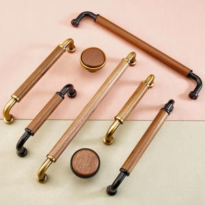 May include: A collection of cabinet hardware, including pulls and knobs. The pulls have a brown wood grain center with gold or black accents. The knobs are round with a matching wood grain finish. The hardware is arranged on a pink and beige surface.