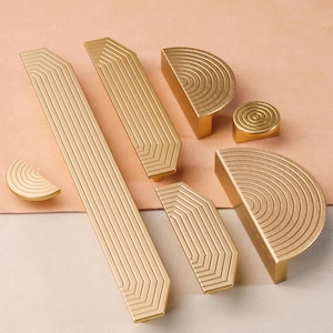 May include: A set of seven gold-toned cabinet hardware pulls with geometric and circular designs. The pulls are made of metal and have a polished finish.