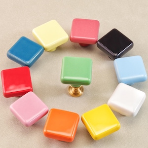 Op de afbeelding: Een verzameling vierkante kastknoppen in verschillende kleuren, waaronder blauw, geel, rood, roze en groen. De knoppen zijn in een cirkelvormig patroon gerangschikt, met een centrale groene knop op een messing basis.