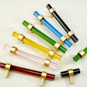 Op de afbeelding: Een verzameling kleurrijke kastgrepen in verschillende tinten, waaronder roodbruin, roze, geel, groen, blauw en zwart. Elke greep heeft een cilindrische handgreep met goudkleurige metalen accenten en een bijpassende basis.