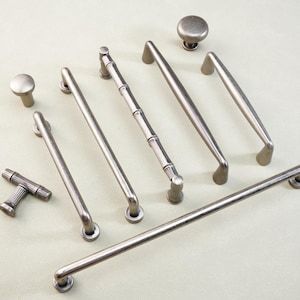 May include: An assortment of antique silver cabinet hardware, including knobs and various handle styles. The collection features a bamboo-style handle, along with several straight and curved bar pulls. The hardware is displayed on a light-colored surface.