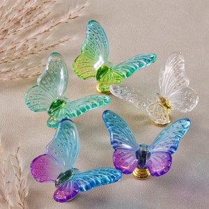 Pode incluir: Uma coleção de figuras de borboletas de vidro coloridas. As borboletas apresentam asas translúcidas em tons de azul, verde e transparente, com bases douradas. As asas têm padrões detalhados.