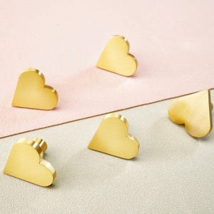 May include: Five heart-shaped cabinet knobs in a warm gold finish are arranged on a pink and grey surface. The knobs are solid and have a smooth, rounded design, perfect for adding a touch of elegance to furniture.