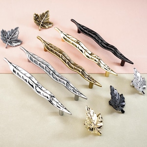 May include: A collection of decorative leaf-shaped cabinet hardware in various metallic finishes, including silver, gold, and black. The hardware includes both pulls and knobs, ideal for adding a natural touch to furniture. The background is a soft pink and beige.
