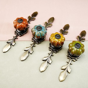 May include: Four decorative cabinet pulls with floral-shaped ceramic knobs in shades of orange, teal, yellow, and green. Each pull has a bronze-colored metal base with a leaf design. The pulls are arranged diagonally against a two-tone background.