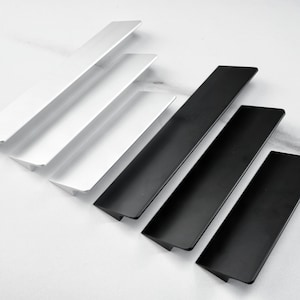 Könnte beinhalten: Eine Sammlung moderner Schrankgriffe in Weiß und Schwarz. Die Griffe sind rechteckig mit abgeschrägter Kante und diagonal angeordnet. Die Hardware ist für Schubladen und Schränke konzipiert.