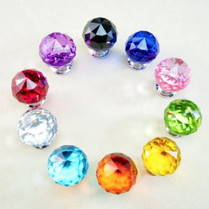 Op de afbeelding: Een verzameling van tien facetgeslepen kristallen kastknoppen in verschillende kleuren, waaronder rood, paars, blauw en groen. Elke knop heeft een zilveren basis en is in een cirkelvorm gerangschikt.